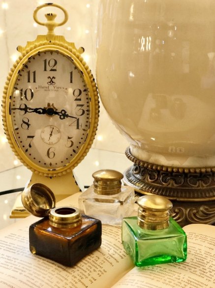 Reloj dorado y tinteros de vidrio: Reloj de sobremesa ovalado en dorado junto a tinteros de vidrio ámbar, transparente y verde.